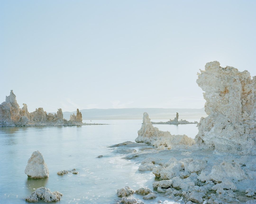 Tufa rock formations rising from the tranquil waters of Mono Lake, California, bathed in soft, diffused light and clear blue sky.