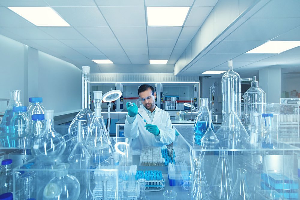 Man in lab coat using pipette in a bright laboratory filled with beakers and scientific glassware, conducting medical research.