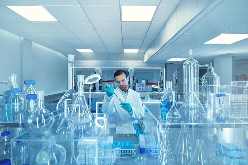 Man in lab coat using pipette in a bright laboratory filled with beakers and scientific glassware, conducting medical research.