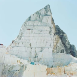 Massive white marble mountain quarry with terraced cuts and small construction machinery at the base against a pale blue sky