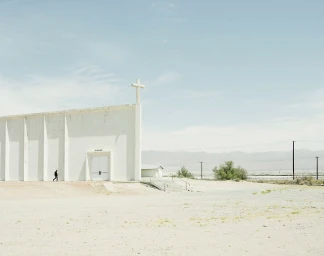 A small figure walking beside a large white concrete church in an arid landscape under a pale blue sky, photographed by Nick Meek.