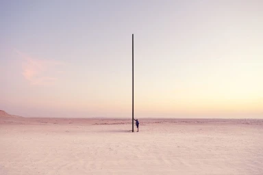 A solitary figure stands beside a towering tall vertical pole in a vast open desert landscape under a pale sunset sky.