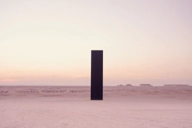 A tall black rectangular monolith stands upright in the middle of a vast flat desert under a pale gradient sunset sky.