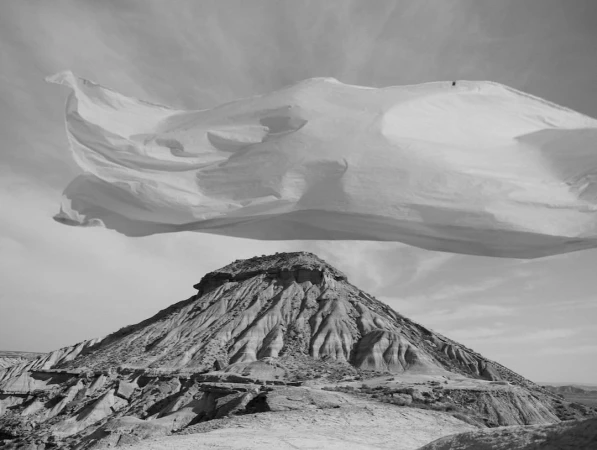 Black and white photograph of a large white fabric sheet floating above an arid mountain landscape by Nick Meek