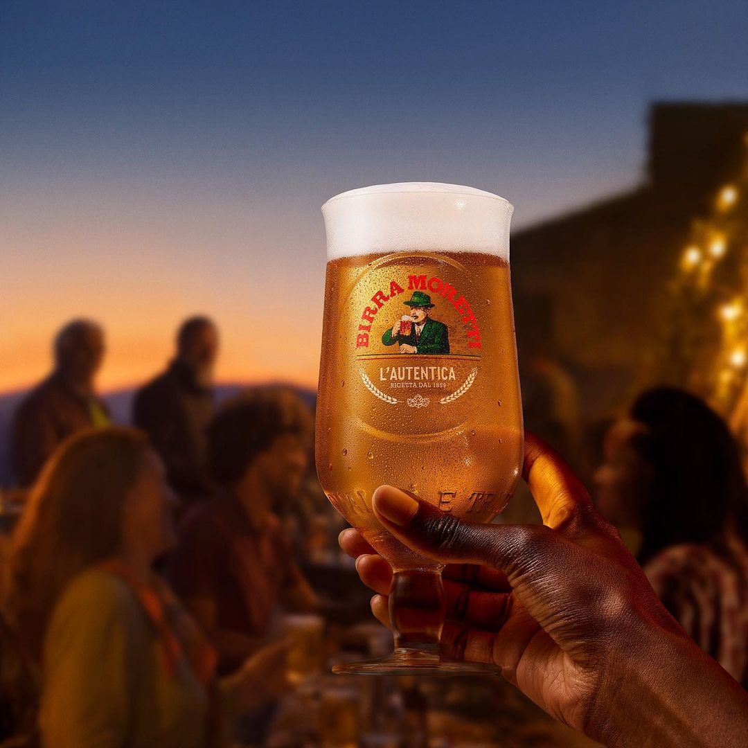 A hand holds a Birra Moretti glass in front of a blurred outdoor gathering at dusk, showcasing the beer and social atmosphere.