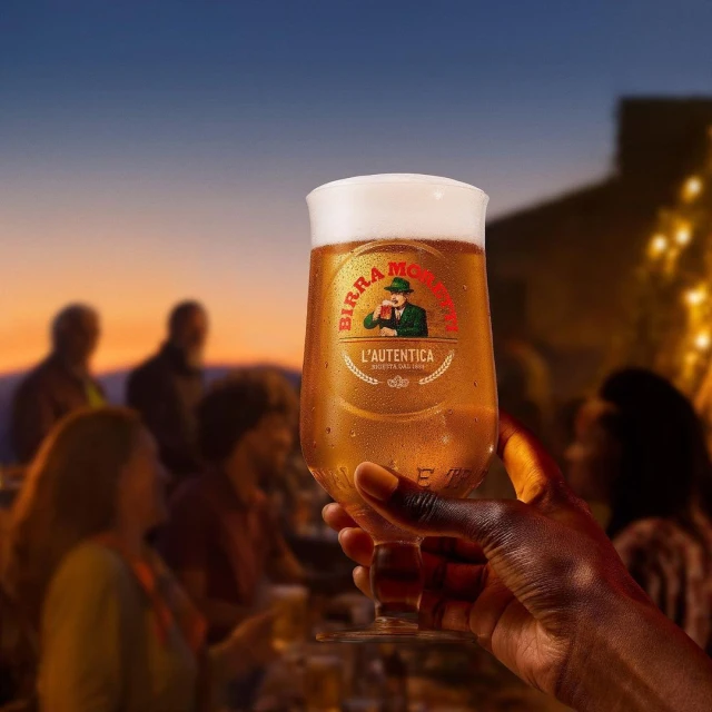 A hand holds a Birra Moretti glass in front of a blurred outdoor gathering at dusk, showcasing the beer and social atmosphere.