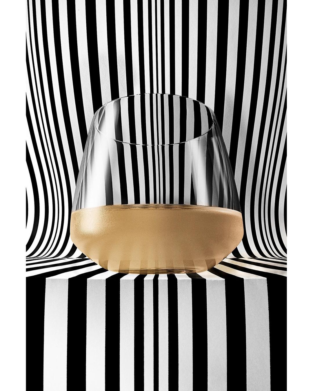 Studio shot of a stemless wine glass filled with a pale golden liquid, set against a striking black and white striped backdrop.