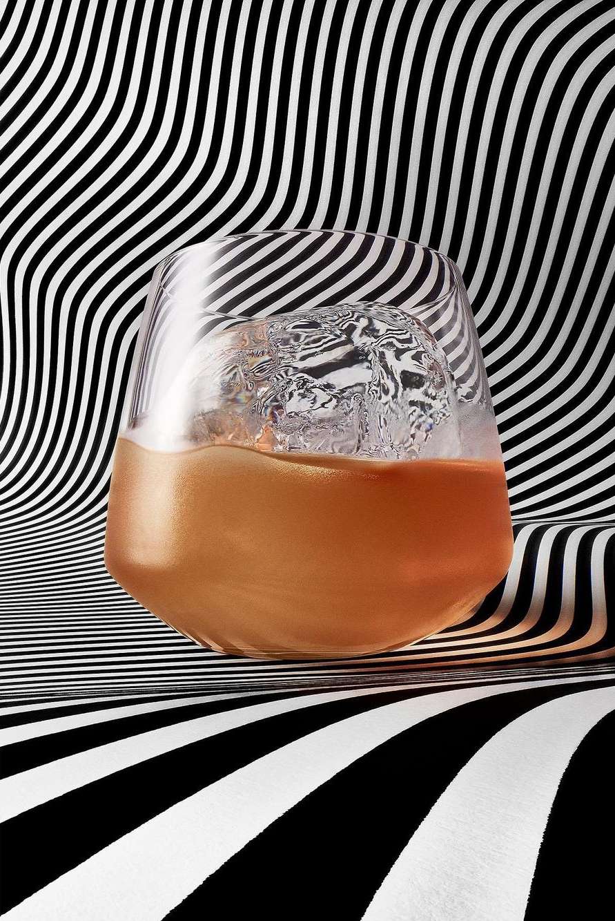 Close-up of a glass filled with a golden liquid and an ice ball, set against a backdrop of bold black and white wavy stripes.