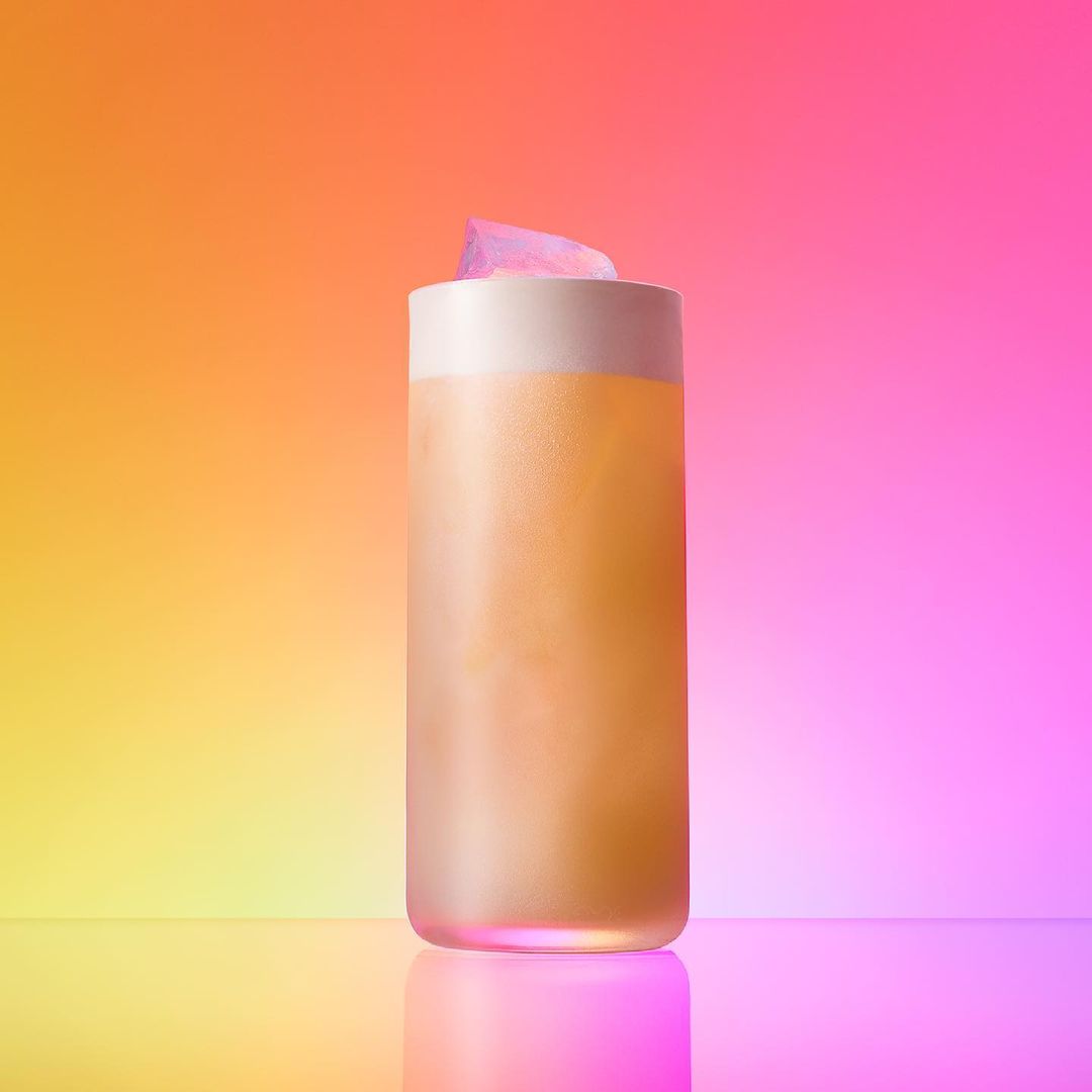 Tall glass filled with a yellow liquid and white foam sits on a reflective surface with a colorful gradient background. Refreshing drink.