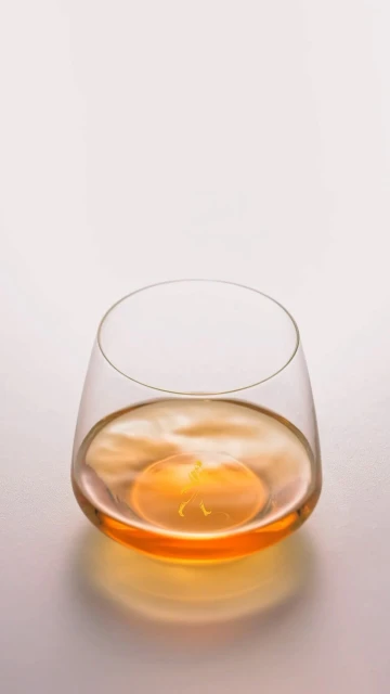 Stemless glass filled with amber liquid, featuring a smooth surface reflecting the light, all set against a plain backdrop.