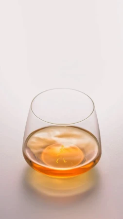Stemless glass filled with amber liquid, featuring a smooth surface reflecting the light, all set against a plain backdrop.