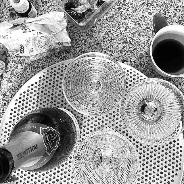 Overhead shot captures champagne bottle and three glasses on a perforated metal tray, accompanied by a cup, snack container, and food packaging.