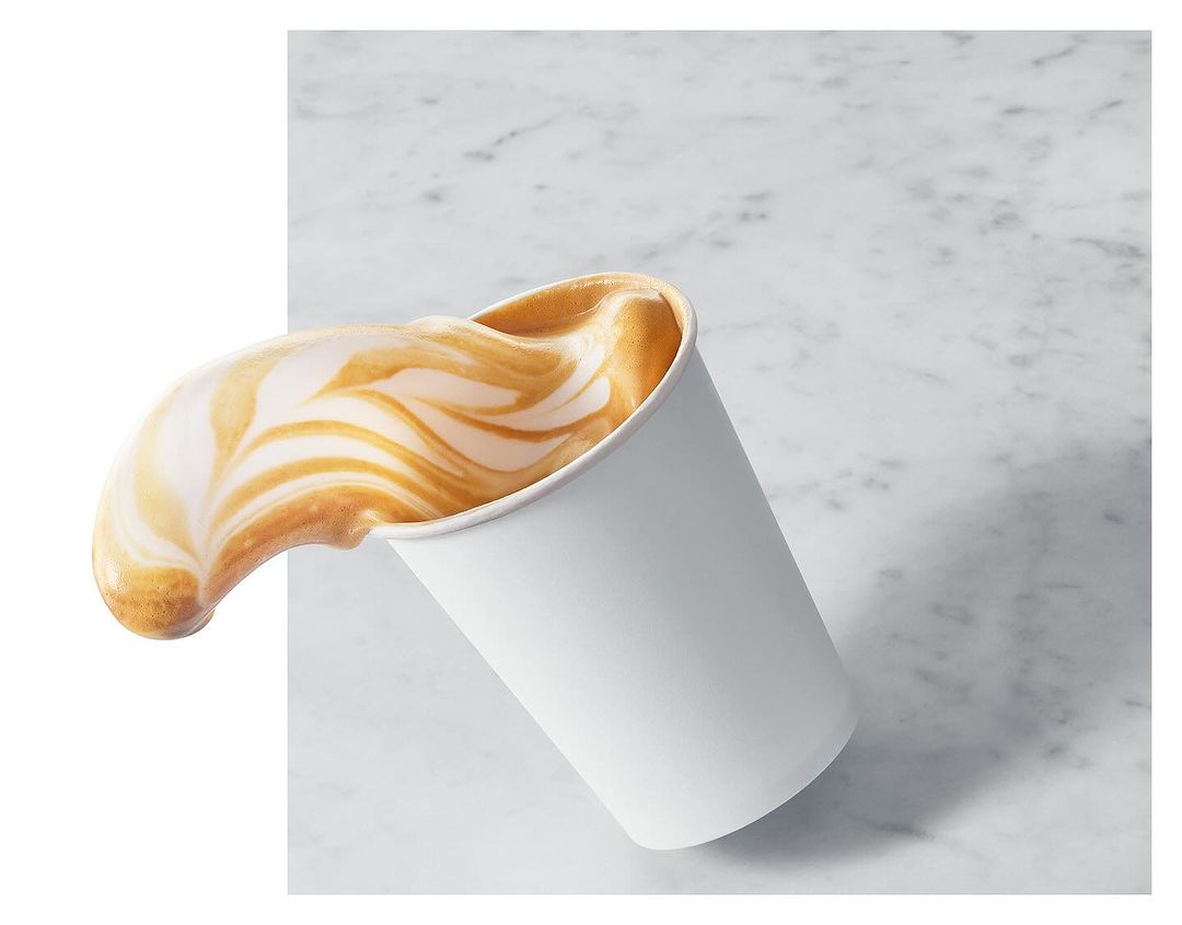 Elevated studio shot of a white paper coffee cup with latte art, foam spilling out against a marble surface and white backdrop.