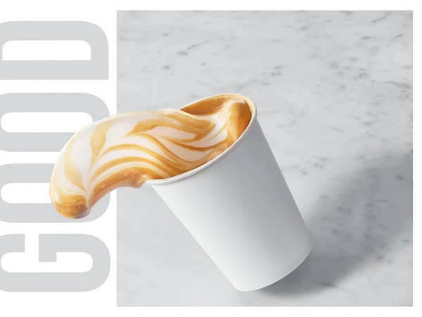 Close-up studio shot of latte art spilling from a tilted white paper coffee cup on a marble surface with light gray text.