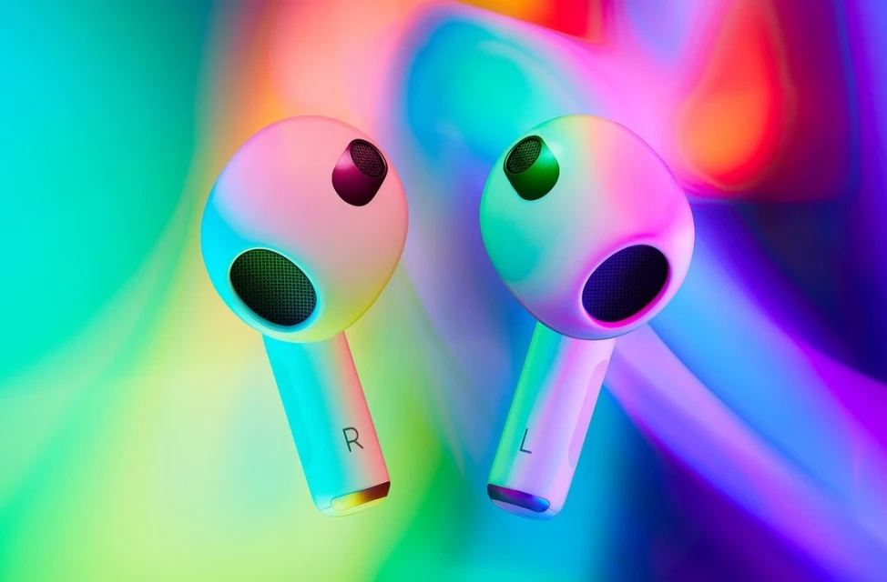 Close-up studio shot of two white wireless earbuds, labeled L and R, set against a vibrant backdrop of blurred turquoise and purple hues.