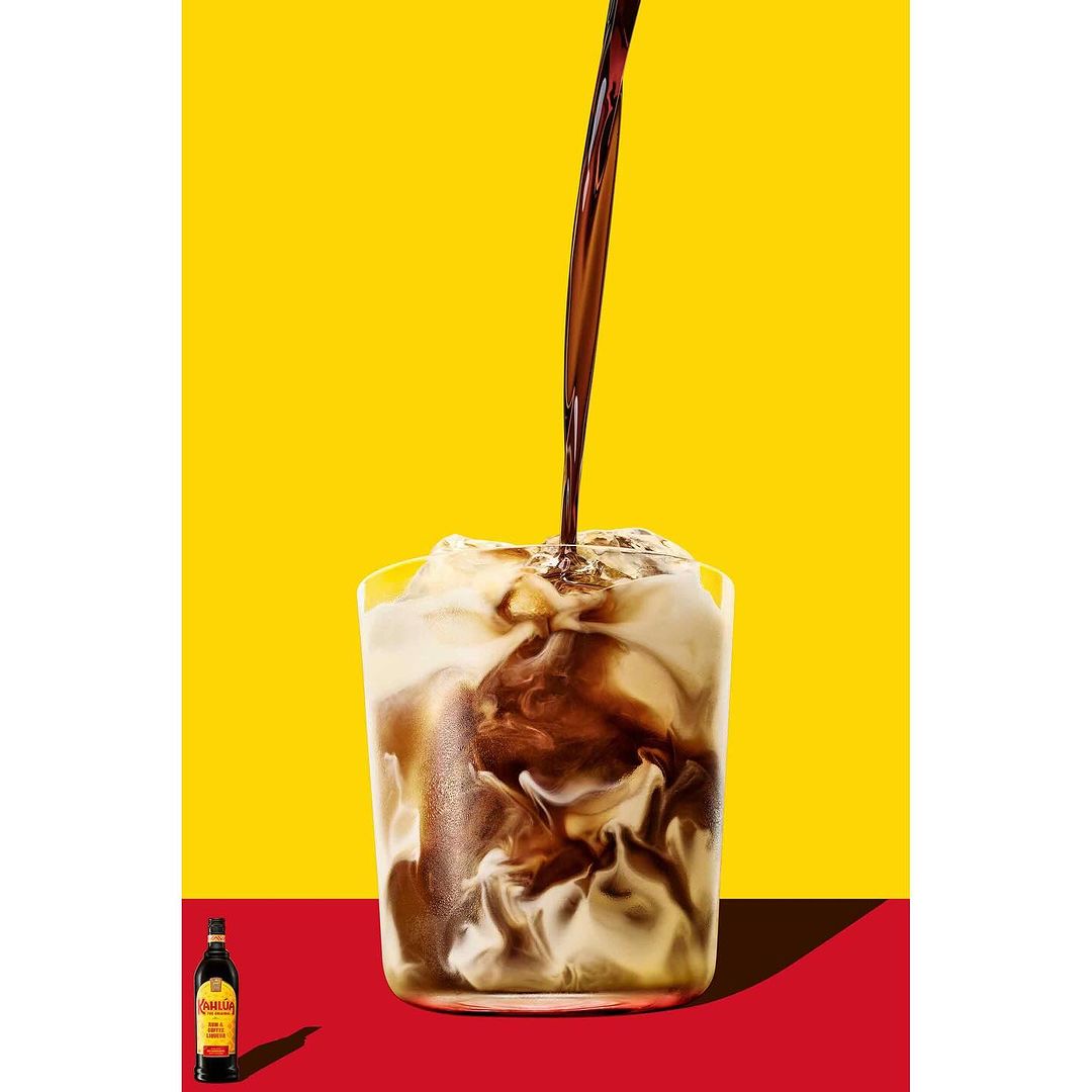 Close-up studio shot of dark liquid pouring into a glass filled with a light-colored beverage against a yellow and red background.