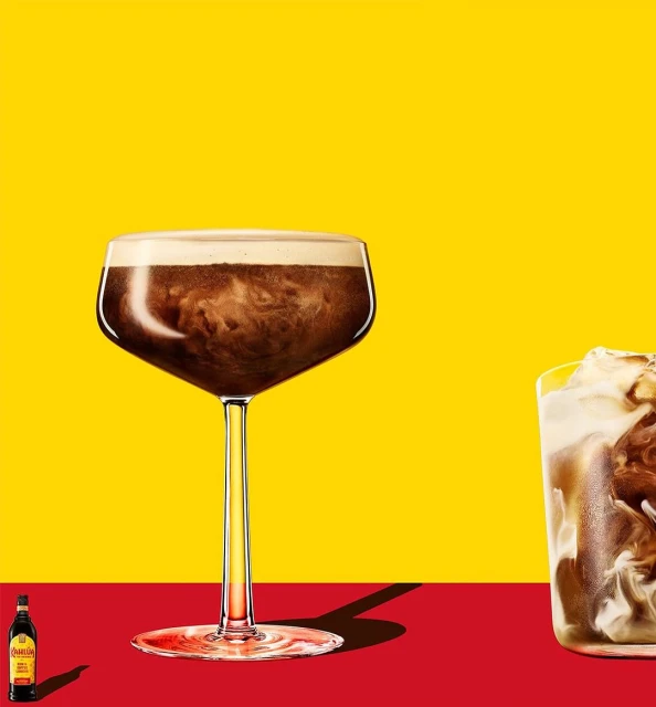 Tall glass of a brown cocktail with white foam on a red table against a yellow background, next to a small bottle.