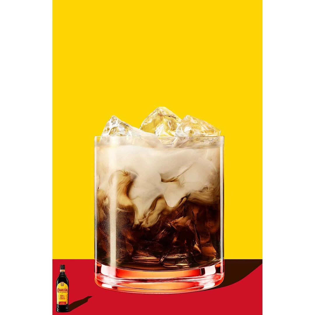 Refreshing White Russian cocktail in glass with ice cubes against vibrant yellow and red background, bottle visible at bottom.
