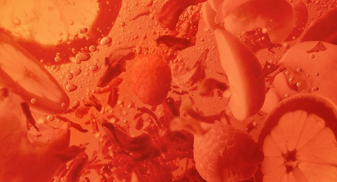 An orange-toned close-up photo of sliced lemons, raspberries, and scattered flower petals suspended in clear liquid with tiny bubbles.