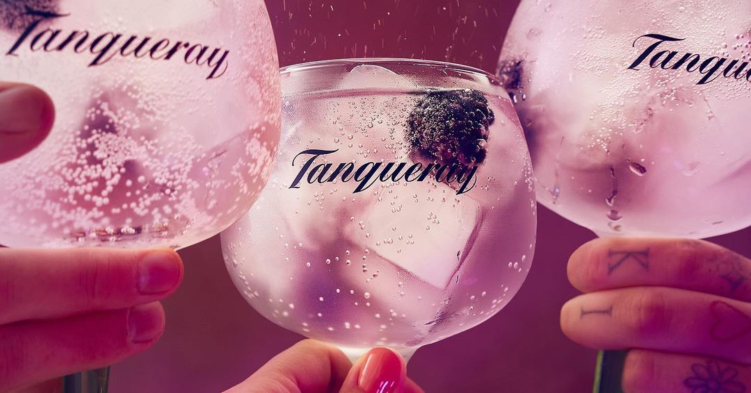 Three clear cocktail glasses filled with ice, clear liquid, and blackberries, held by two people against a pink background with logo.