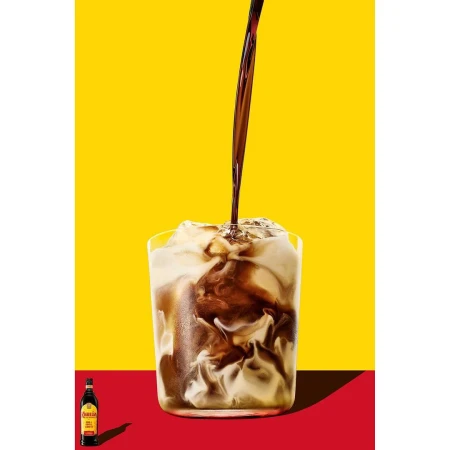 Dark liquid pouring into a glass of ice and milk against a bright yellow and red background by photographer Nick Rees