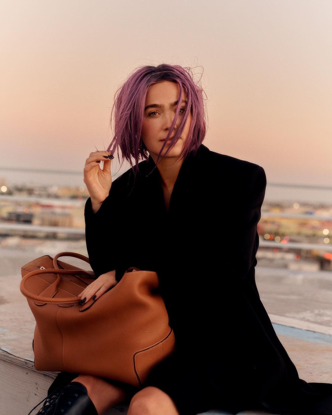Woman with purple hair sits on rooftop with a black coat and a tan leather tote bag at golden hour, city buildings blurred.