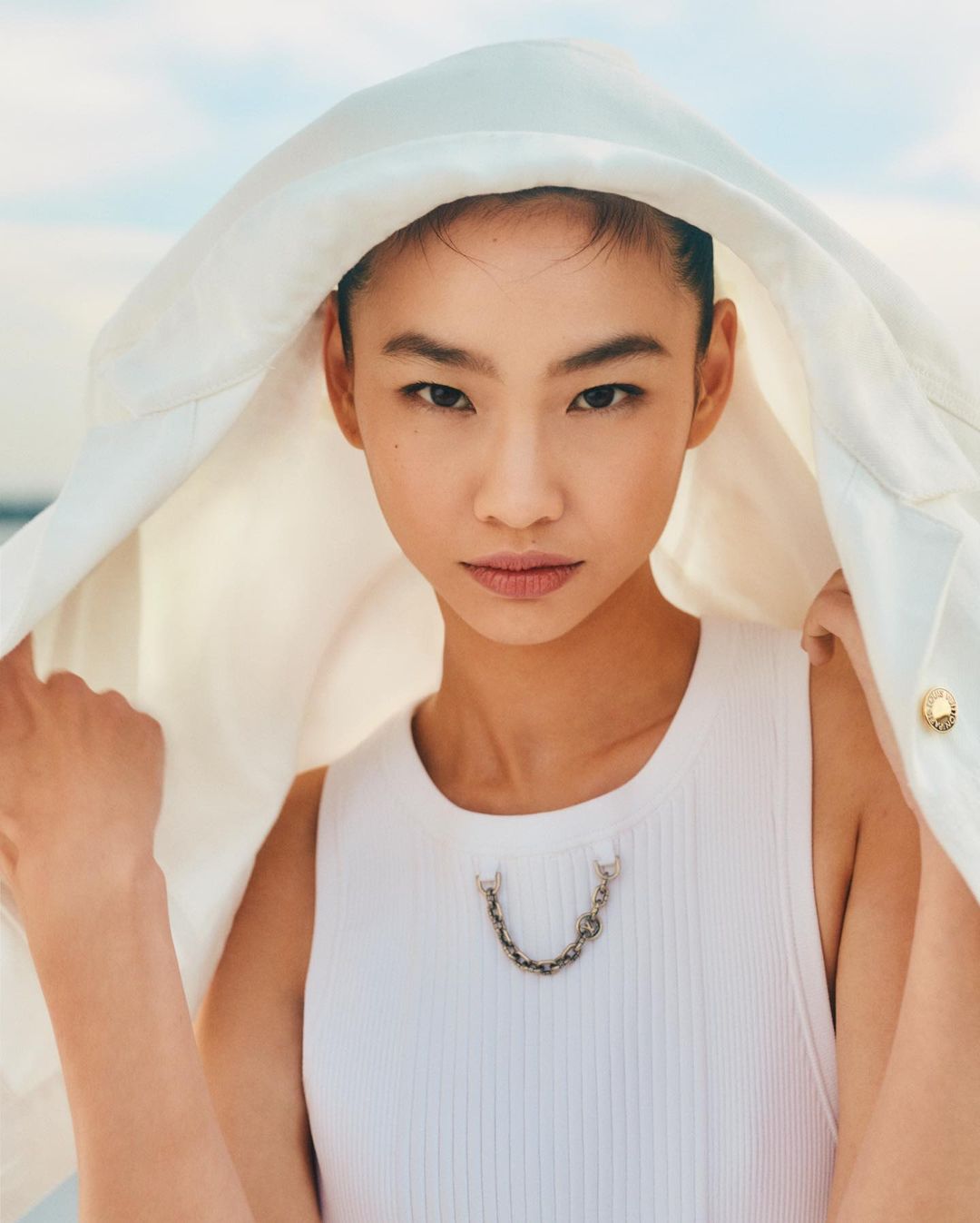Portrait of a young Asian woman wearing a white sleeveless top and jacket with hood against a blurred background of sky and water.