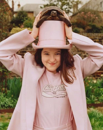 Smiling woman poses outdoors in pink suit with a tortoise on her pink hat. A blurred stone wall and green plants form the backdrop.