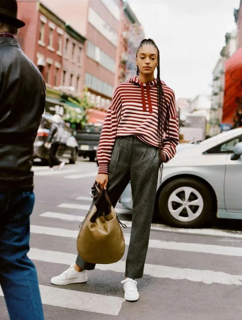 Woman in striped hoodie and grey trousers standing on a crosswalk, holding a tan handbag, in an urban environment, outdoors.