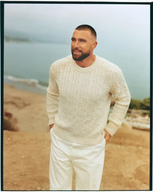 Man in a cream-colored cable knit sweater and white pants stands smiling on a sandy bluff overlooking the ocean on a cloudy day.