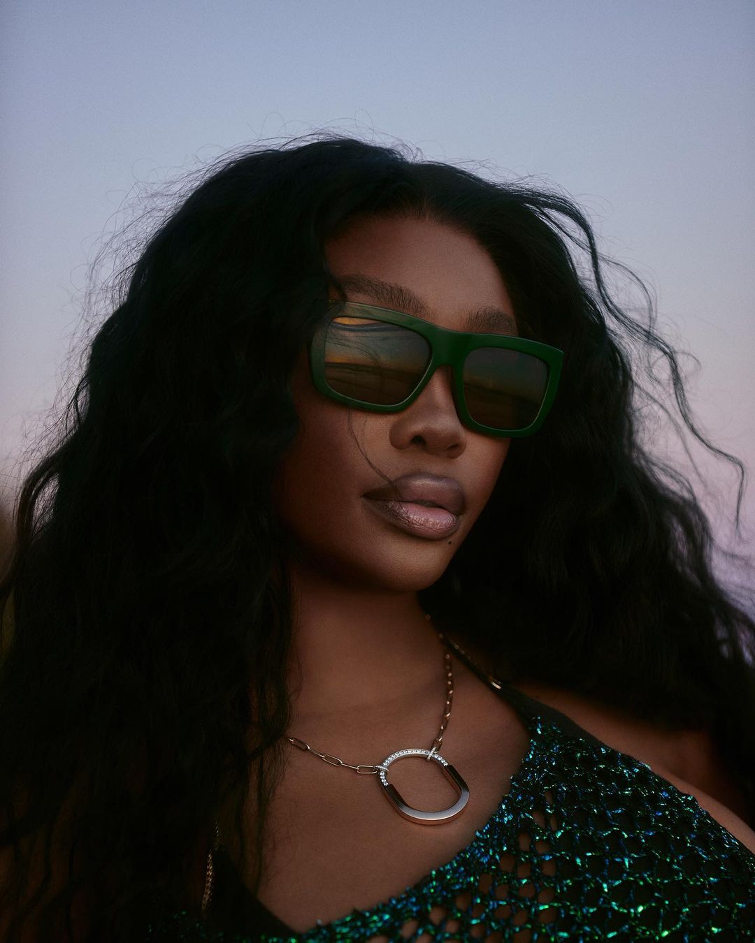 Close-up portrait of a woman with dark hair and green sunglasses, wearing a gold necklace and a green, sparkly top against a dusky sky.