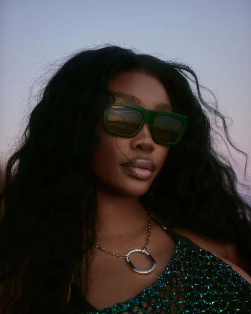Close-up portrait of a woman with dark hair and green sunglasses, wearing a gold necklace and a green, sparkly top against a dusky sky.