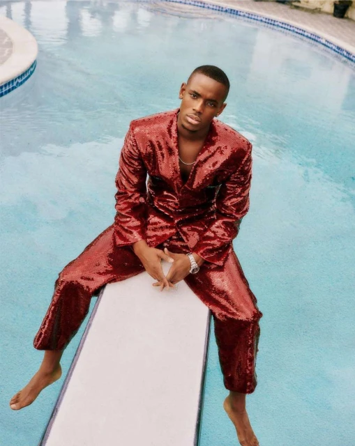 A stylish young black man in a red sequin suit poses on a white diving board next to a bright blue pool in an outdoor setting.