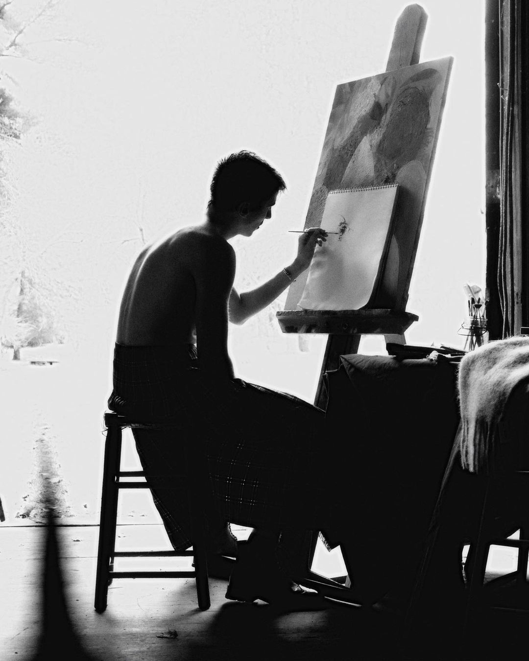 Black and white photo of a shirtless person sitting and painting at an easel in a bright room. Art studio setting with natural light.