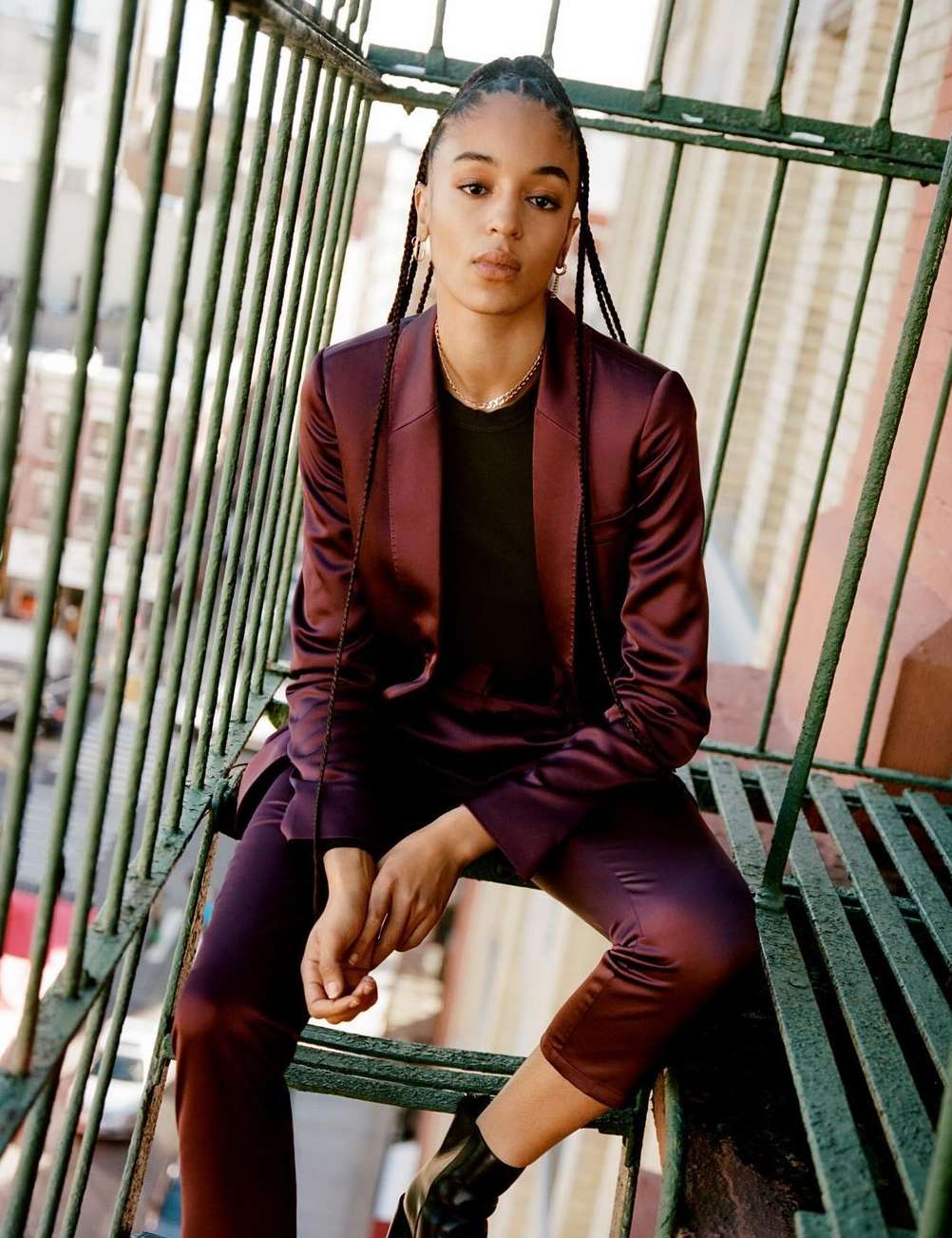 Adult female with braids sits on outdoor stairs wearing a tailored aubergine-colored pantsuit with black top and boots in an urban environment.