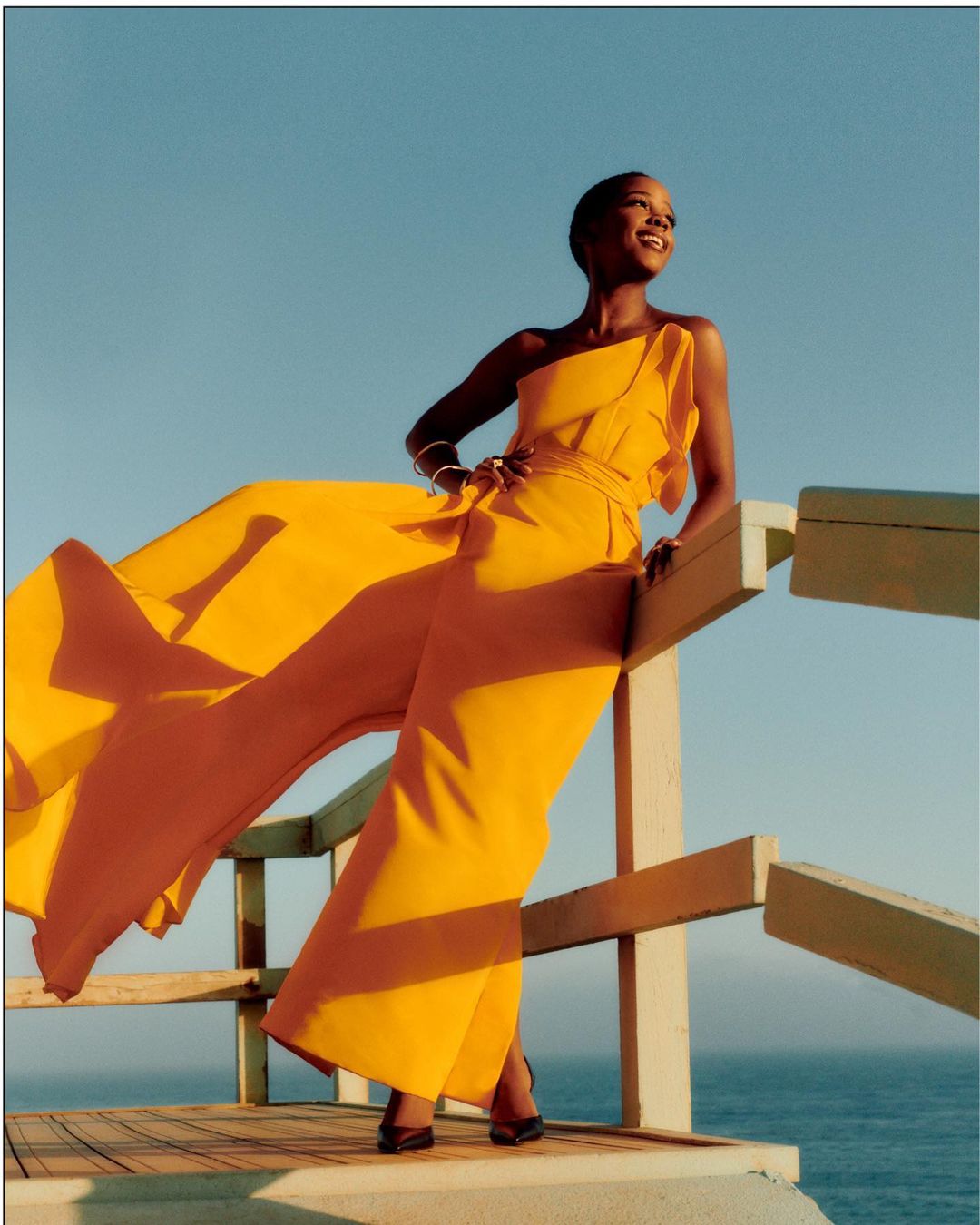 Stylish woman in a yellow jumpsuit smiles and poses against a bright blue sky and ocean backdrop, leaning on a wooden structure.