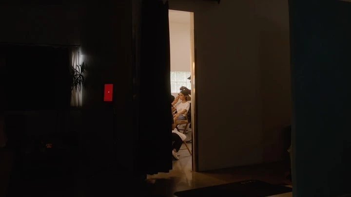 Adult female sits in chair getting her makeup done in a room that's partially visible through doorway with a dark room in the foreground.