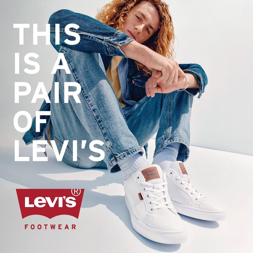 A young adult man with curly red hair poses while seated in jeans and jacket next to a pair of white Levi's sneakers on a white background.