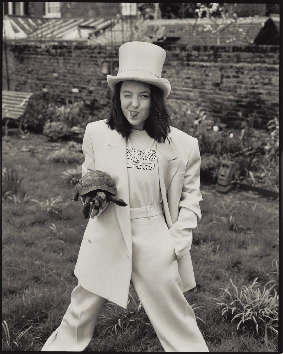 Monochrome portrait of a woman in a suit and top hat, holding a turtle, standing on a lawn with a house in the background.