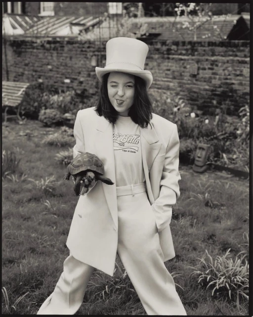 Monochrome portrait of a woman in a suit and top hat, holding a turtle, standing on a lawn with a house in the background.
