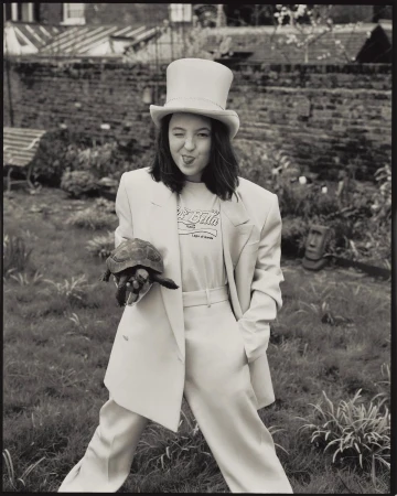 Monochrome portrait of a woman in a suit and top hat, holding a turtle, standing on a lawn with a house in the background.