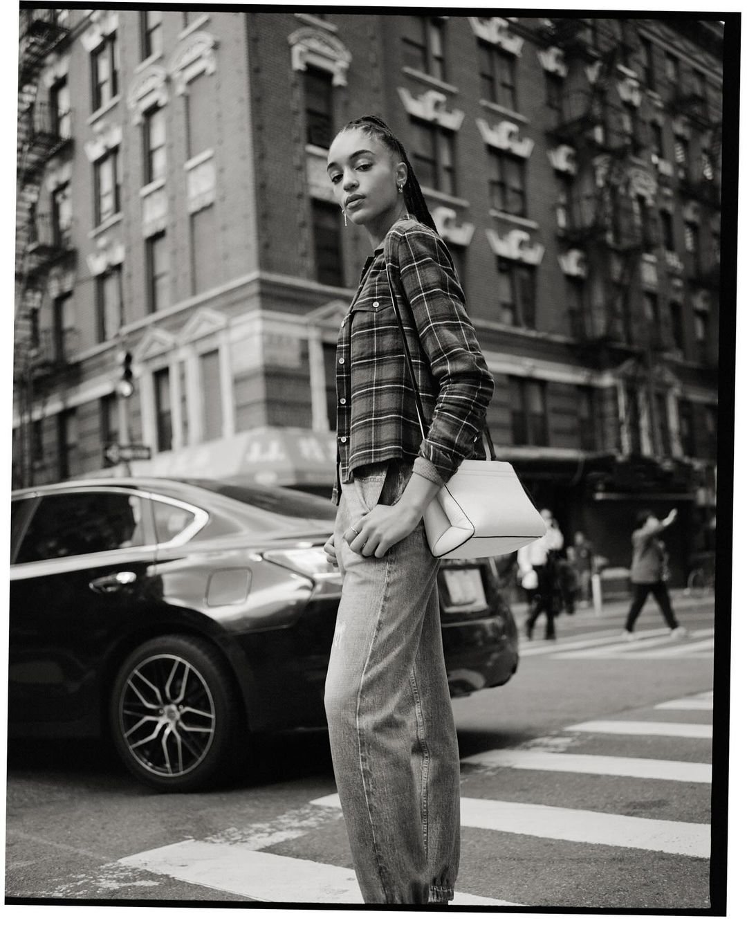 Model with braided hair wearing a plaid shirt and jeans standing near a parked car on a city street.