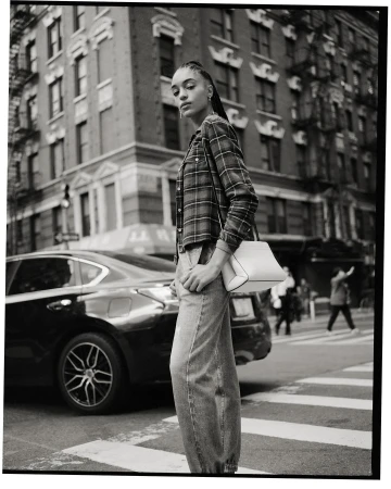 Model with braided hair wearing a plaid shirt and jeans standing near a parked car on a city street.