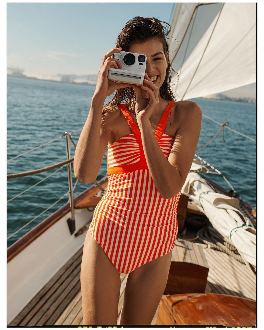 Smiling woman in striped swimsuit takes a photo with an instant camera on a sailboat, with blue water and sky in the background.