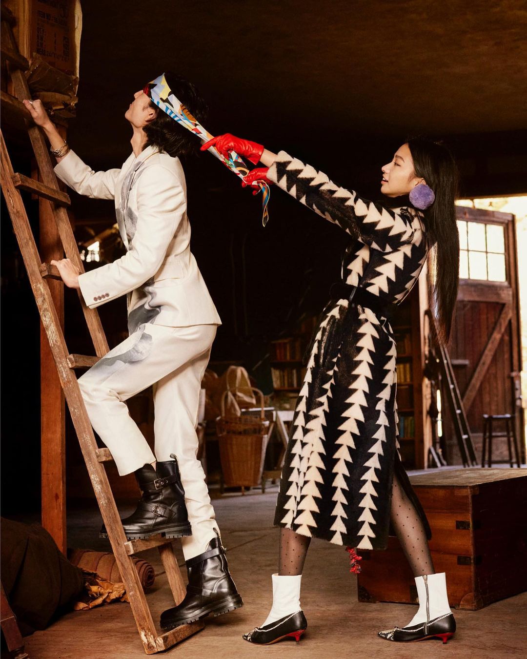 Two fashion models in an indoor setting: a man climbing a wooden ladder in a white suit, and a woman holding a scarf.