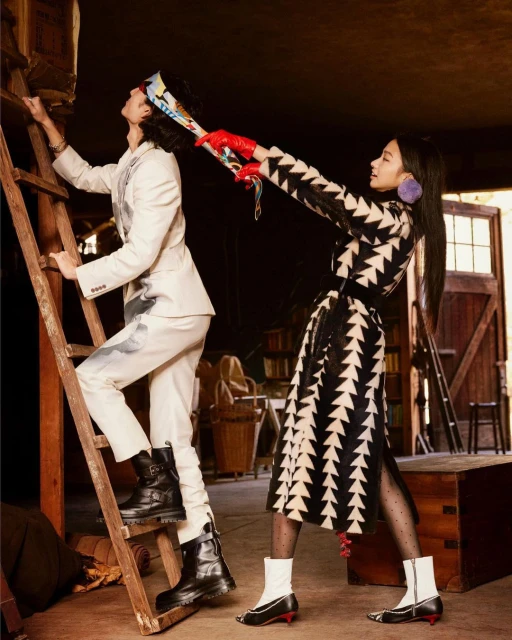 Two fashion models in an indoor setting: a man climbing a wooden ladder in a white suit, and a woman holding a scarf.