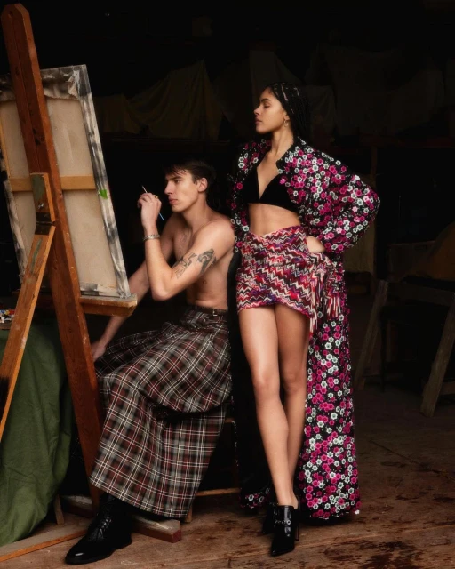 A shirtless man sits at an easel painting while a woman in a floral-patterned robe stands nearby in an art studio with soft, diffused lighting.