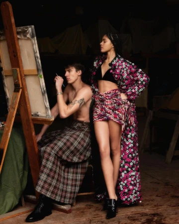 A shirtless man sits at an easel painting while a woman in a floral-patterned robe stands nearby in an art studio with soft, diffused lighting.