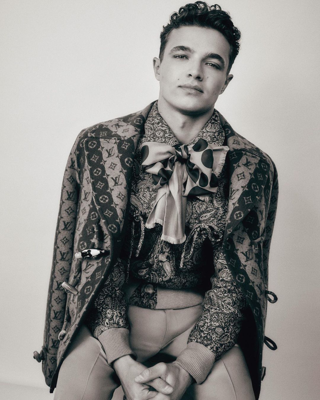 Monochrome portrait of male model with curly hair wearing patterned jacket and shirt with bow tie in a studio setting.
