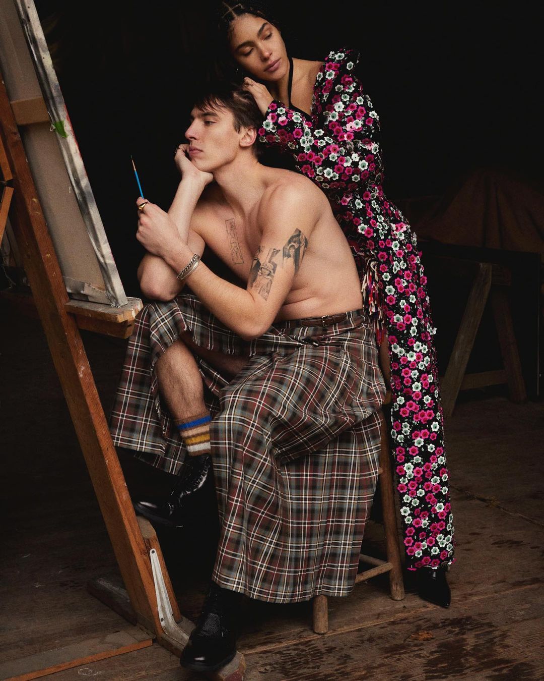 A man in a kilt sits by an easel with a painting, while a woman in a floral dress stands behind him in a dimly lit indoor setting.